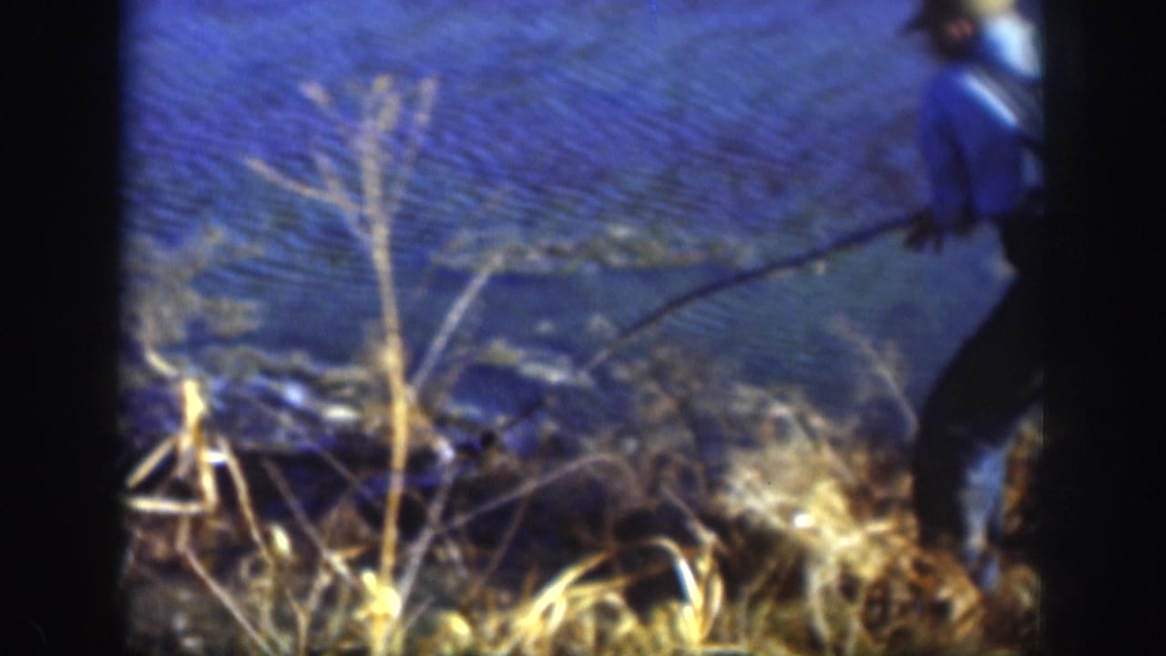 Man in blue jacket at the right edge of frame, partially cropped, holding a fishing pole at a lake shoreline with dry grass in the foreground.