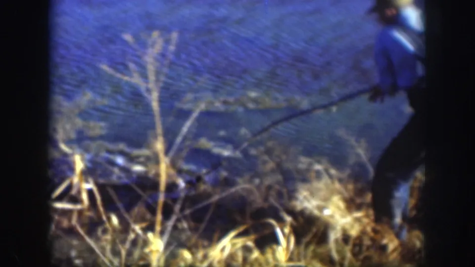 8mm home movie frame showing a man in a blue jacket with a fishing pole at a rural lake shoreline, likely Kansas, circa 1958.