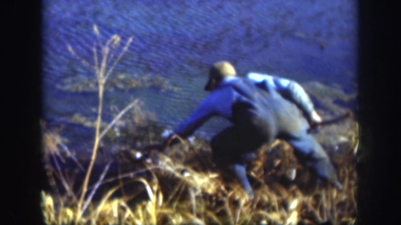 8mm home movie frame showing a man in a blue jacket bent low at a rural lake shoreline amid dry golden grass, consistent with retrieving a catch, Kansas circa 1958.