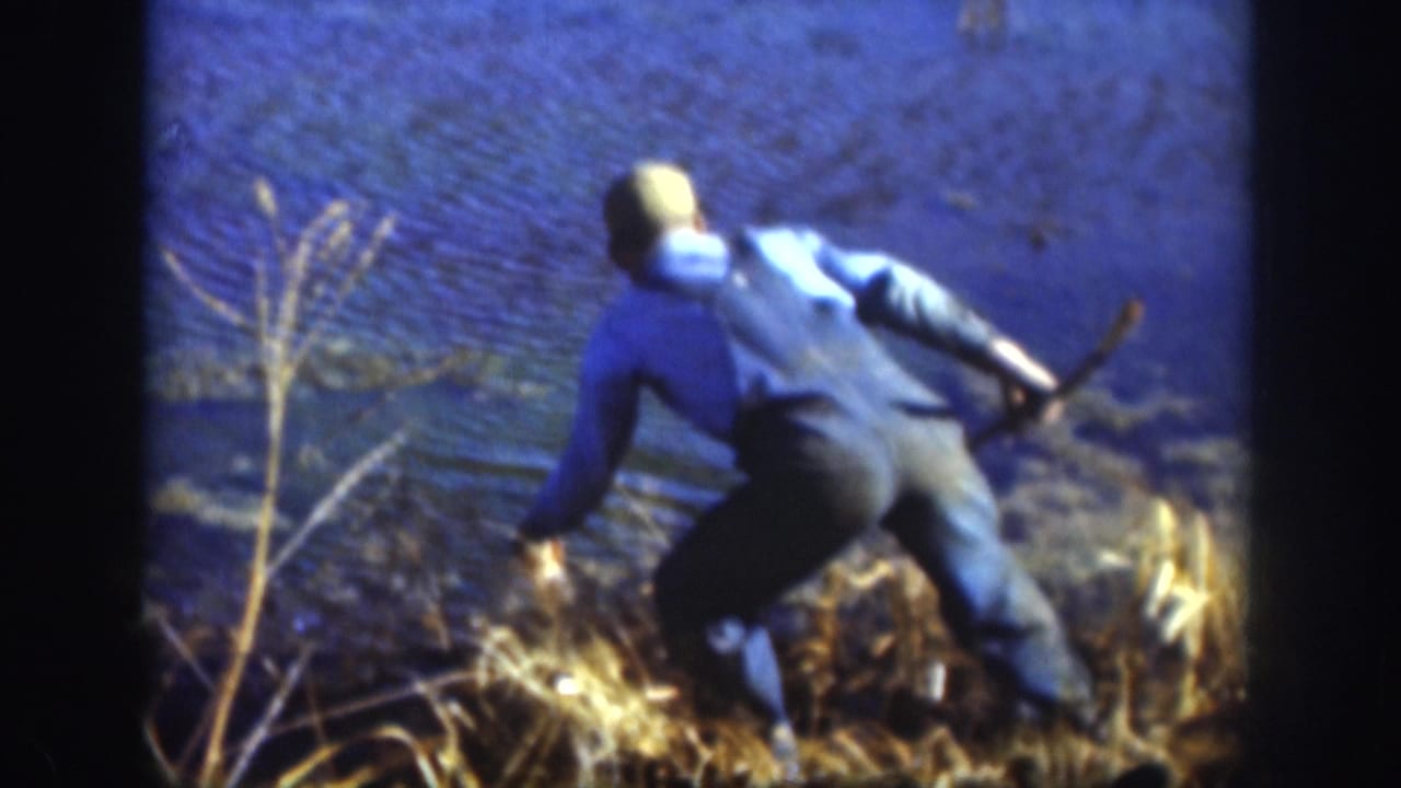 8mm home movie frame showing a man in a blue jacket bent low at a rural lake shoreline amid dry grass, likely handling a fish, Kansas circa 1958.