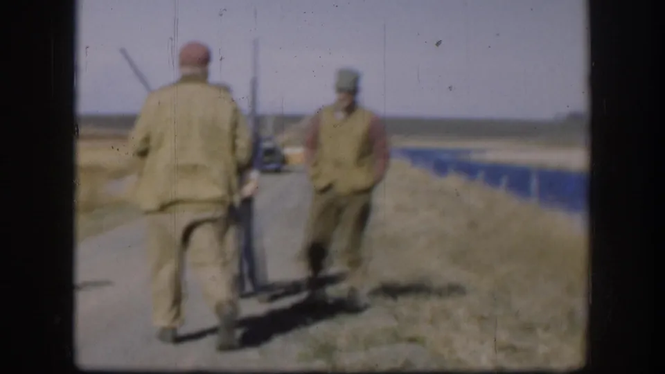Two men in hunting attire walk away from camera along a flat Great Plains road, rifle visible, utility poles in background, circa 1958.