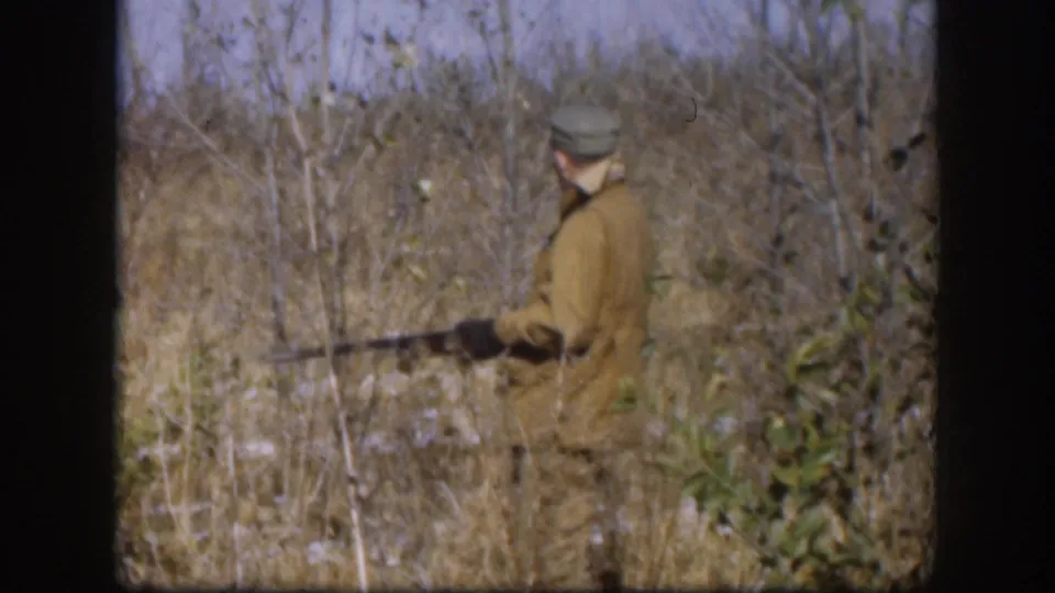 Hunter in tan coat and cap carrying a rifle through dry brush, Kansas, circa 1958; leafless shrubs and pale winter sky visible in background.