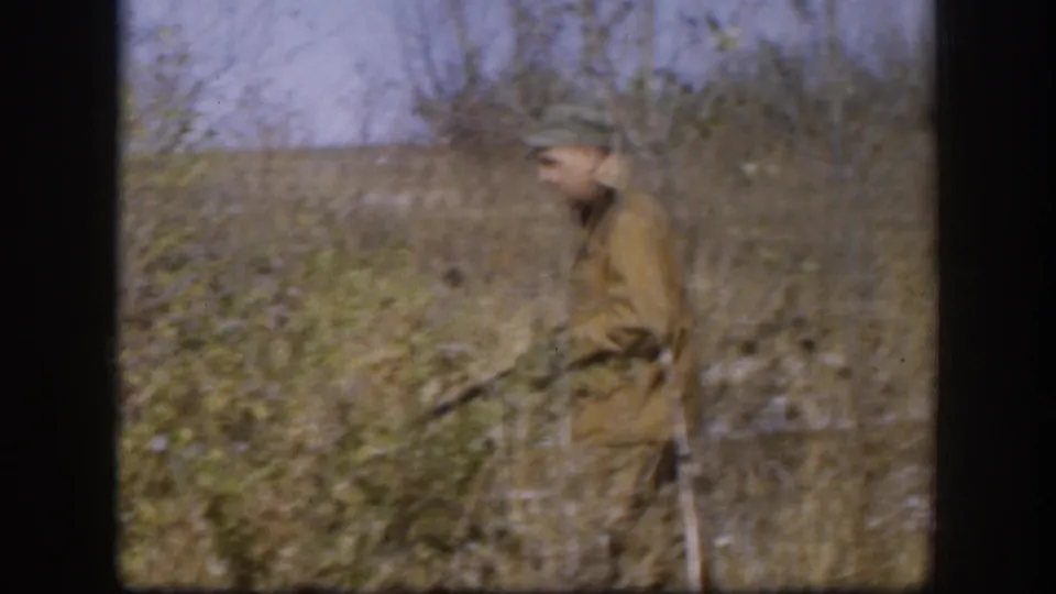 Hunter in tan coat and cap pausing in a dry Kansas brush field, circa 1958; leafless shrubs and pale overcast sky visible in background.