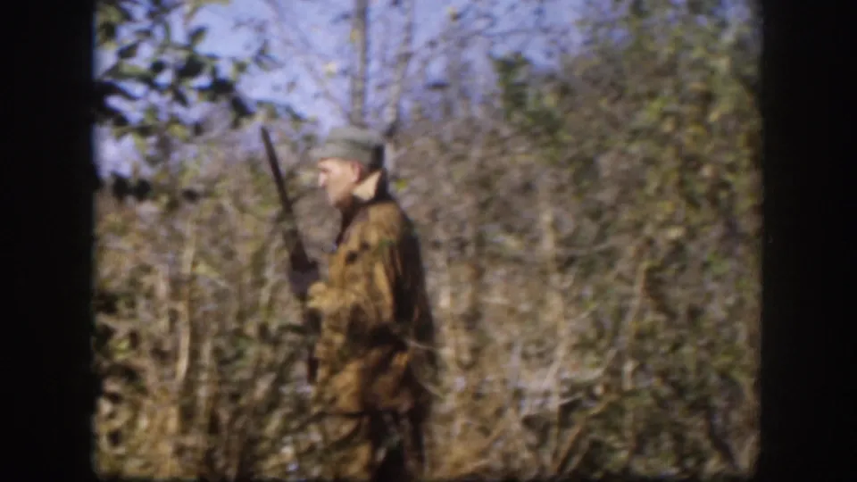Hunter in camouflage jacket and pale cap carries a long-barreled firearm upright among brush and bare trees, 8mm home movie, Kansas circa 1958.