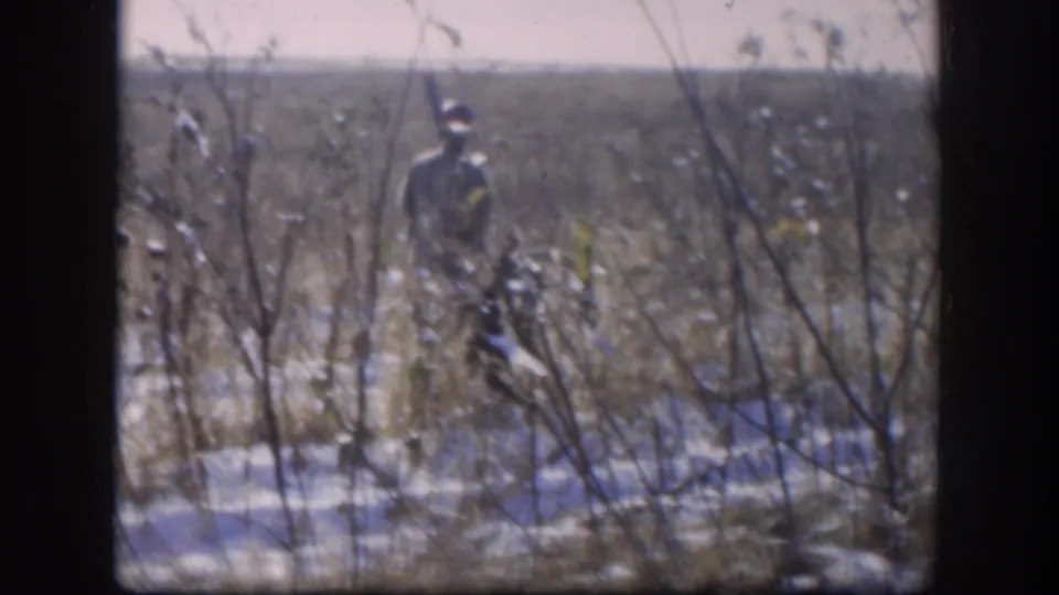 1958 Kansas 8mm home movie: hunter seen from inside through a window, moving through winter scrub brush with snow on the ground and bare branches in the foreground.