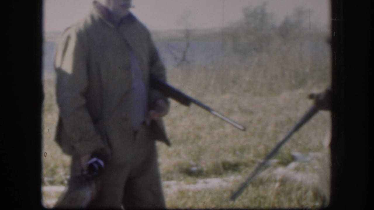Close crop of a hunter in a tan jacket gripping a rifle at his side in an open field, from a Kansas 8mm home movie circa 1958.