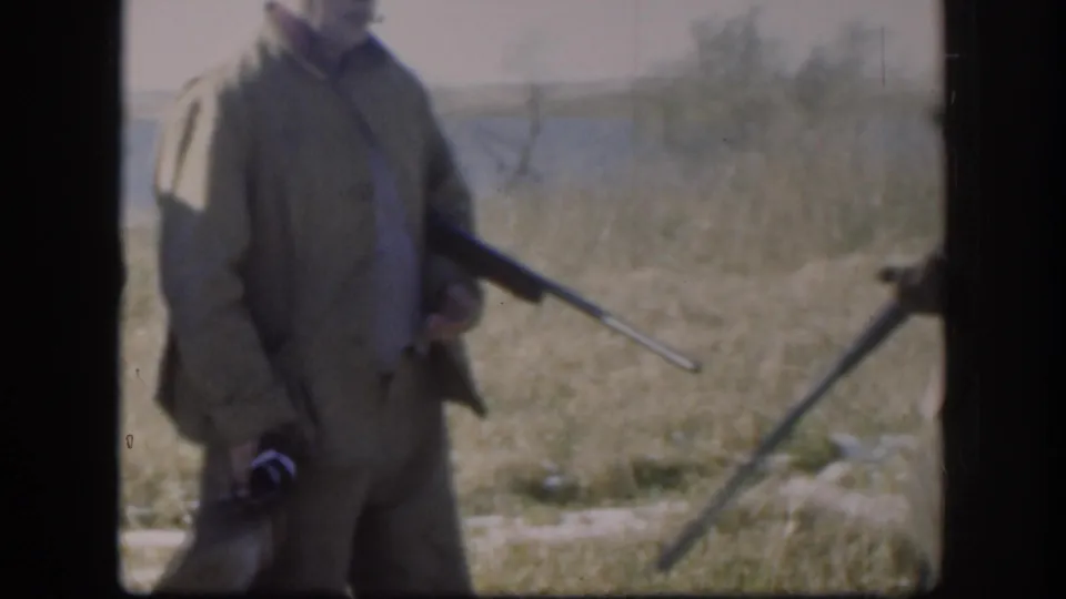 Close crop of a hunter in a tan jacket gripping a rifle at his side in an open field, from a Kansas 8mm home movie circa 1958.