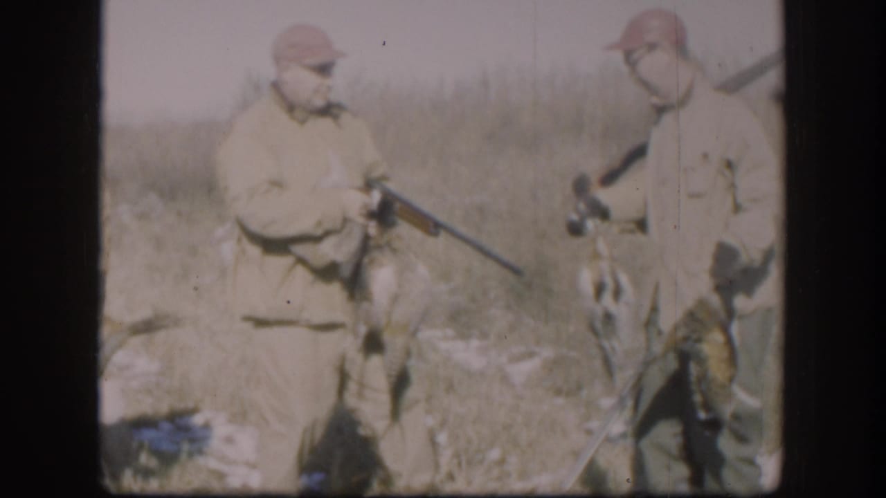 Two men in tan jackets and caps standing in a field, one holding a rifle; dark shapes near their feet.