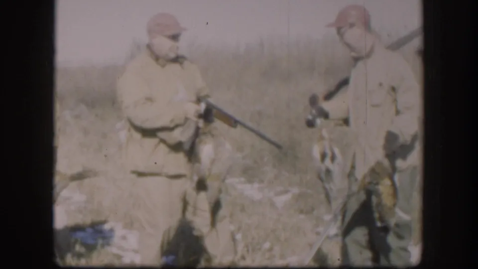 Two hunters in tan jackets and caps pose in a Kansas field with a rifle and what appear to be game birds at their feet, circa 1958 8mm home movie.