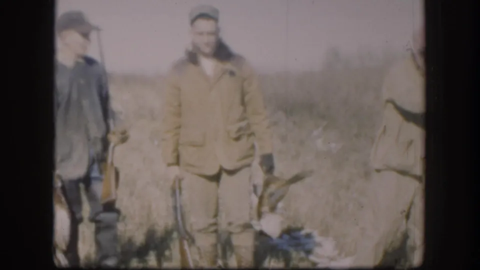 Two hunters in tan jackets stand in an open Kansas field with harvested game birds at their feet, from a 1958 8mm home movie.