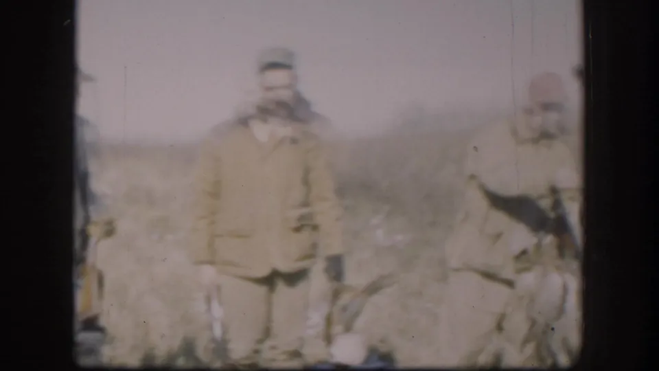 A lone hunter in a tan jacket stands in a Kansas field with what appear to be game birds at his feet, from a 1958 8mm home movie.