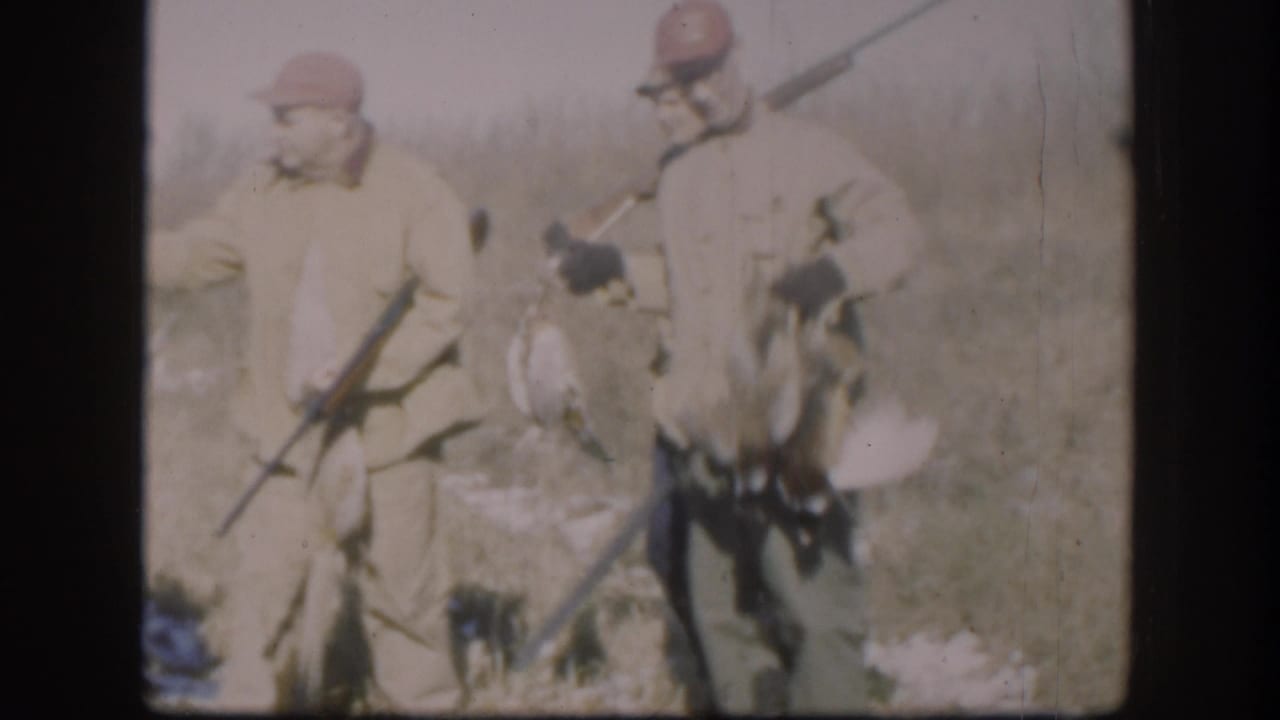 Two hunters hold harvested birds aloft while a third stands with a shotgun in a flat rural field with snow patches.