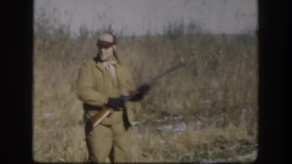 A lone hunter in 1950s-era tan hunting jacket and orange-red cap holds a shotgun in dry Kansas grassland with snow on the ground, circa 1958.