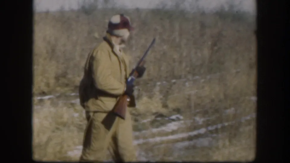 A hunter in profile view wearing a tan jacket and orange-red cap carries a shotgun across a snow-dusted Kansas field, circa 1958.
