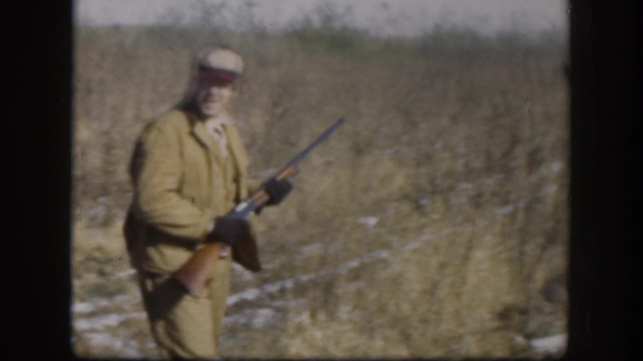 Adult male hunter in tan jacket and light cap holds a shotgun while walking through a dry grass field with snow patches, Kansas circa 1958.