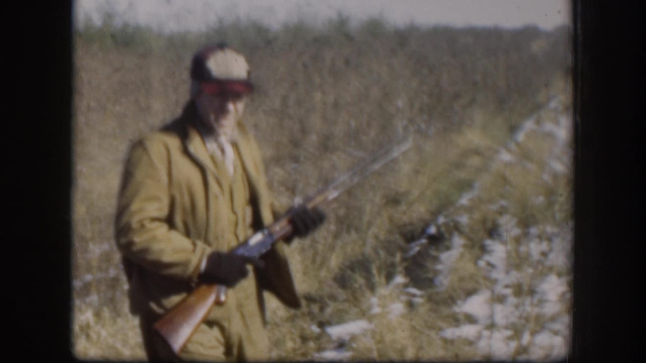 Hunter in tan jacket turned toward camera, holding a break-action shotgun with wooden stock, dry grass and snow visible behind him.