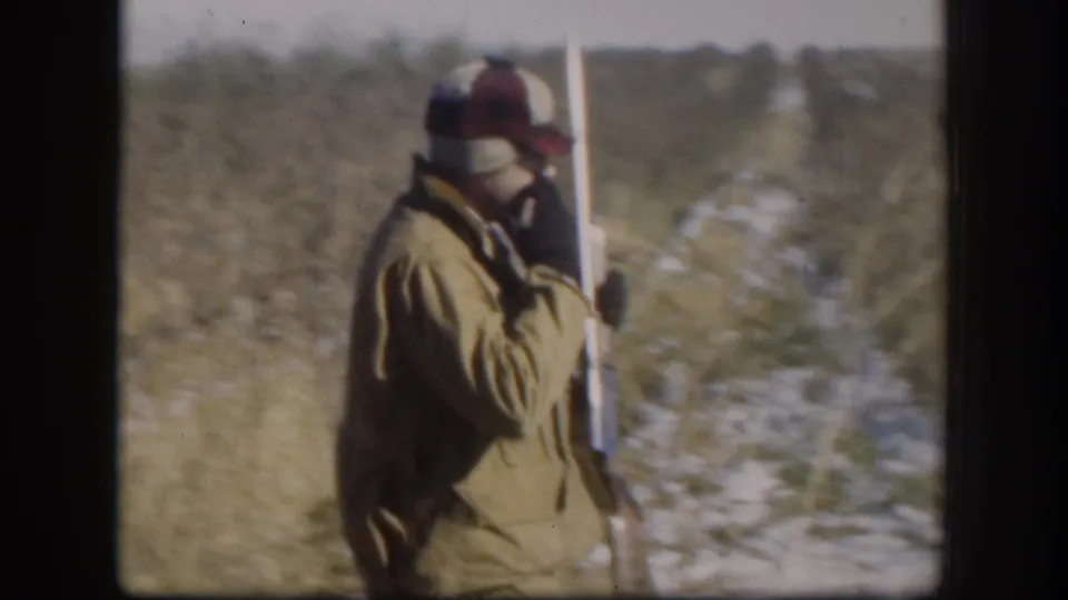 Hunter walking toward camera in tan jacket, shotgun carried upright, snow-patched field stretching behind, Kansas circa 1958.