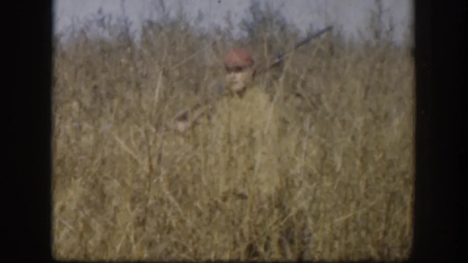 Hunter in tan jacket mostly concealed within tall winter grass, crouching in apparent stalking position in a Kansas field circa 1958.
