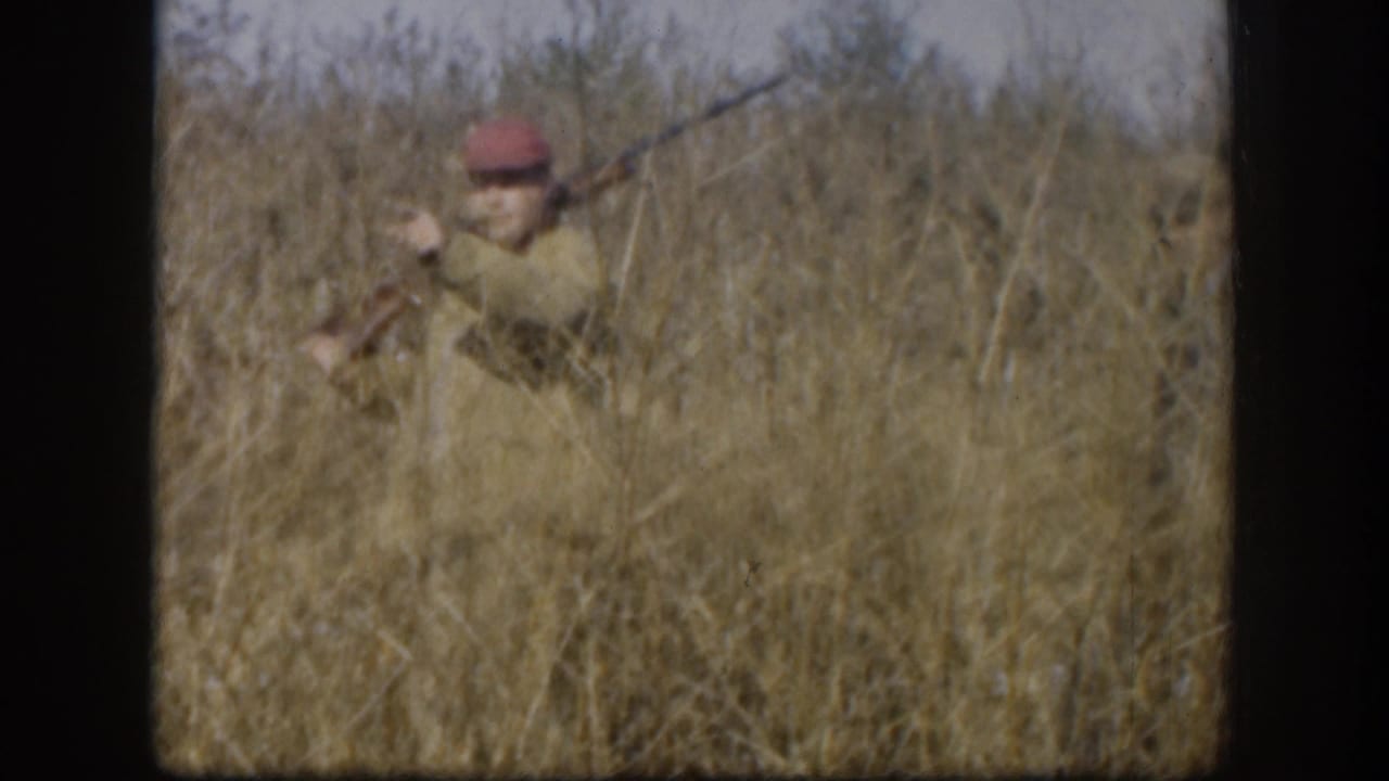 Single hunter in red hunting cap and light tan jacket raises a shotgun amid tall golden-brown grass, consistent with late 1950s Kansas hunting footage.