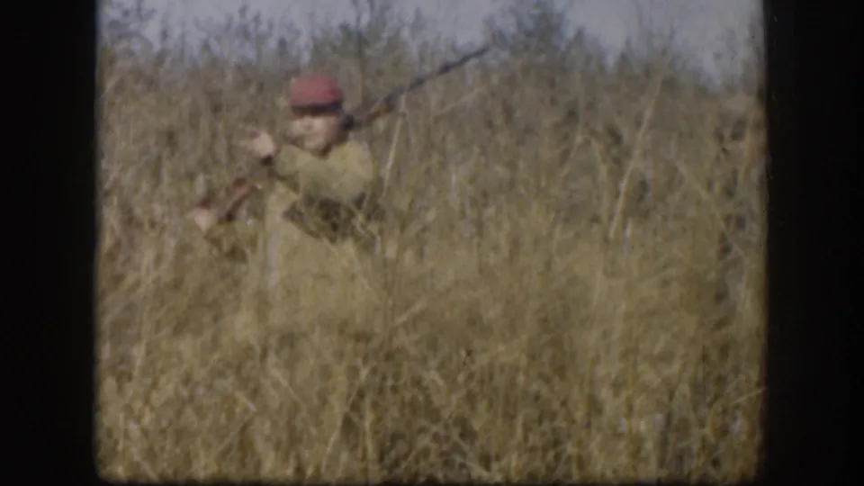 Single hunter in red hunting cap and light tan jacket raises a shotgun amid tall golden-brown grass, consistent with late 1950s Kansas hunting footage.