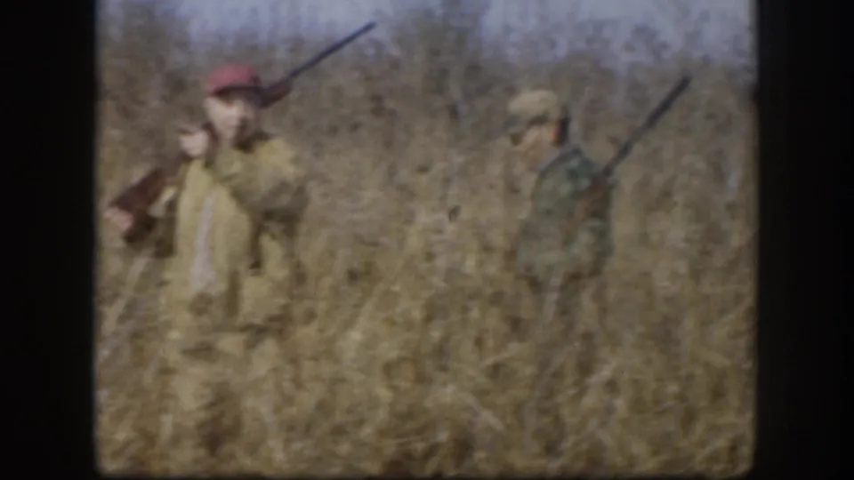 Two hunters with shotguns wade through dry autumn grass; foreground hunter wears a red cap and tan jacket, second a patterned jacket; pale sky and sparse bare brush visible behind.