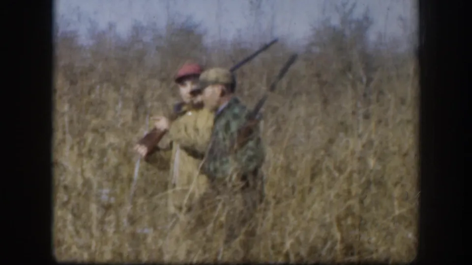 Two hunters in dry tall grass, red-capped man on left and camouflage-jacketed man on right, both with shotguns raised; consistent with late 1950s Kansas rural hunting footage.