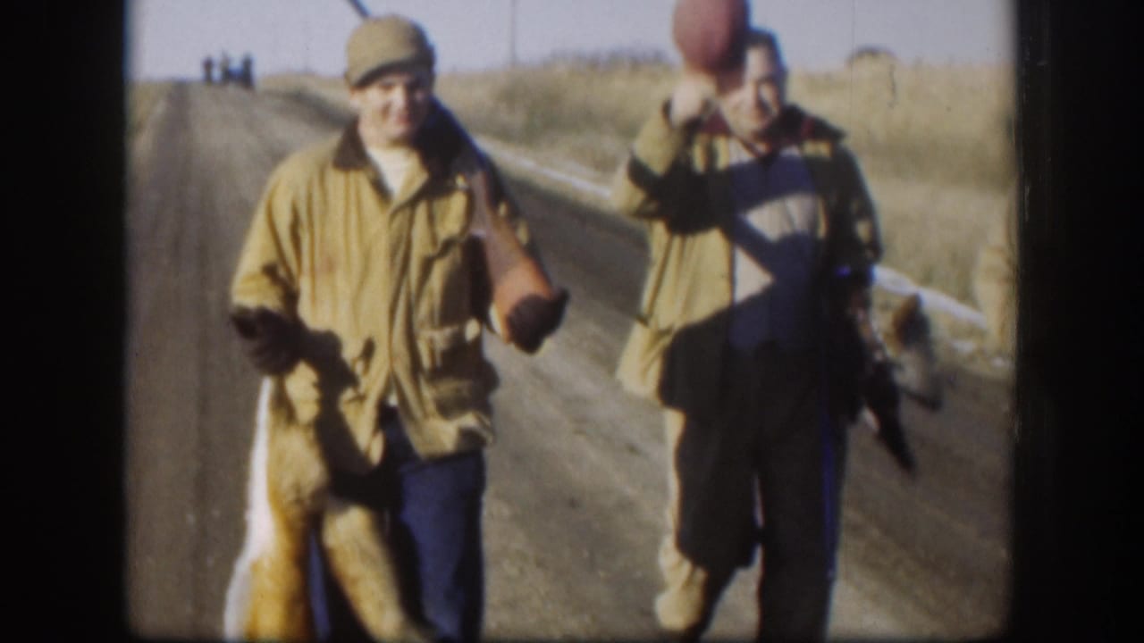 Two hunters in olive field jackets and caps on a Kansas gravel road, c. 1958; one holds a dead fox by the hind legs while his companion looks on.