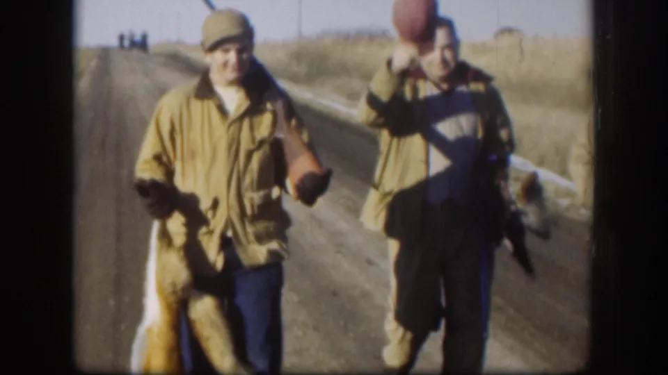 Two hunters in olive field jackets and caps on a Kansas gravel road, c. 1958; one holds a dead fox by the hind legs while his companion looks on.