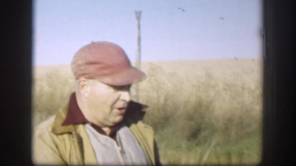 Hunter in red baseball cap and olive field jacket speaking toward camera, c. 1958; flat Kansas prairie, dry grass, and a wooden utility pole visible behind him under clear blue sky.