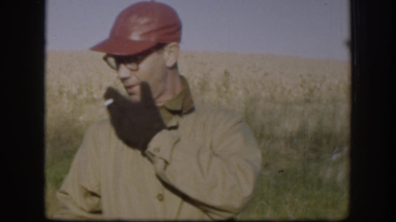 Man in red cap and wire-frame glasses smokes a cigarette, golden wheat field behind him.