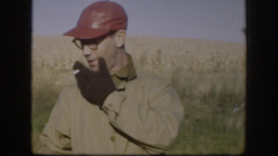 Man wearing red cap, wire-frame glasses, and tan jacket smoking a cigarette in front of a golden wheat field, Kansas, circa 1958, 8mm color.