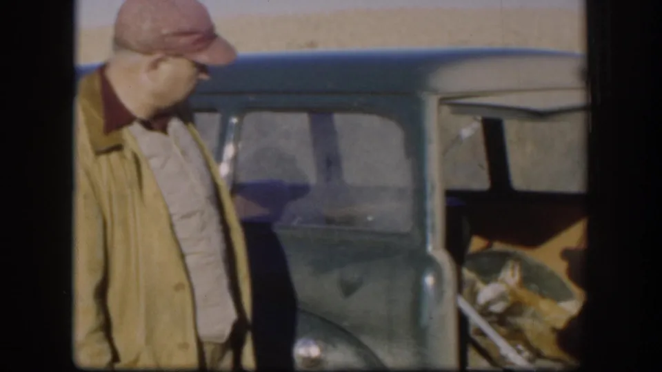 Man in red cap and tan work jacket beside a vintage blue-green vehicle with open door, wheat field horizon behind, Kansas circa 1958, 8mm color film.