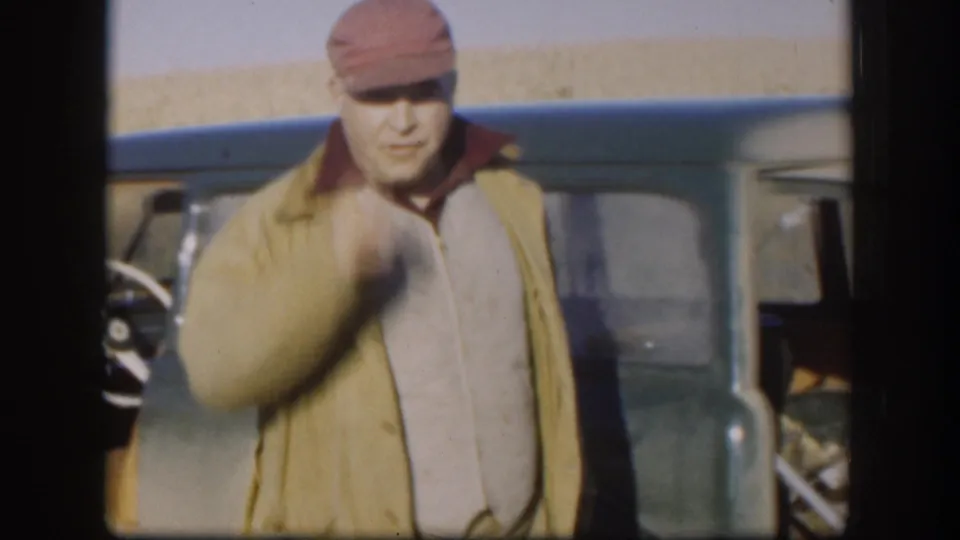 Man in red cap and tan jacket raising his arm beside a vintage blue-green vehicle, Kansas wheat field horizon, circa 1958, 8mm color.