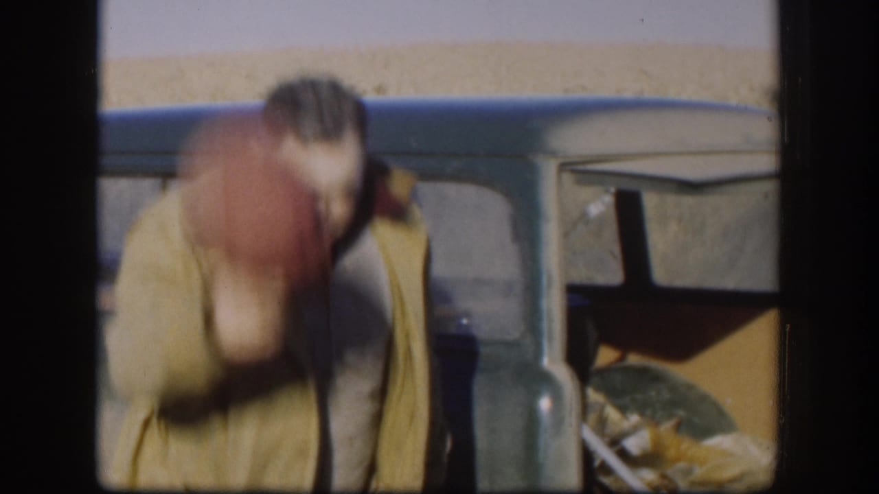 Man in tan jacket leaning forward beside a dark classic car; face motion-blurred; orange-furred animal visible in vehicle cargo area.