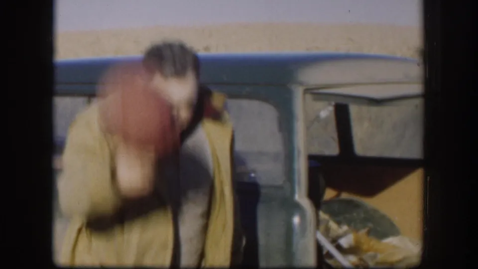 8mm home movie frame circa 1958, Kansas; man in tan outdoor jacket bends toward a dark classic car with a dead fox visible in the open cargo area.