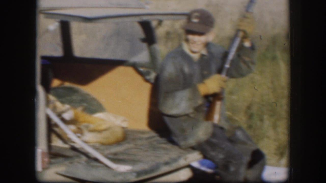 Man in dark jacket and lettered ball cap holds a rifle; dead fox laid on a vehicle's open cargo surface beside him.