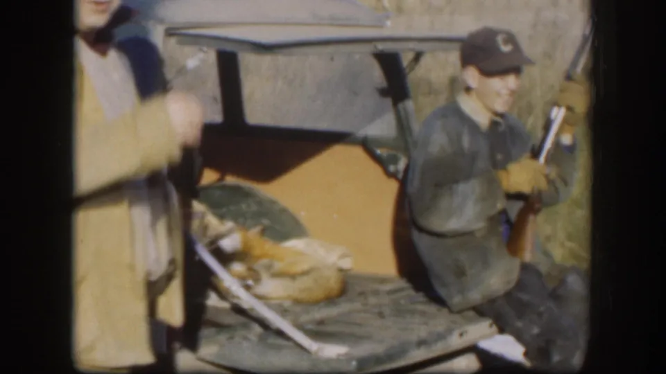8mm home movie frame circa 1958, Kansas; smiling hunter in lettered ball cap holds a rifle beside a dead fox on a vehicle tailgate, with a second hunter partially visible at frame edge.