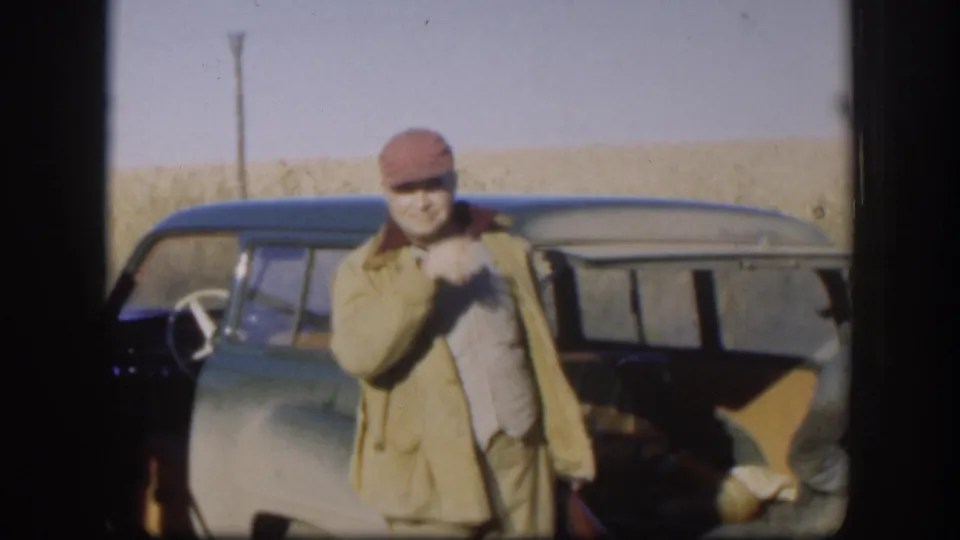 8mm home movie frame circa 1958, Kansas; man in tan jacket and red cap poses smiling in front of a dark mid-century car in open prairie, with a utility pole at right.