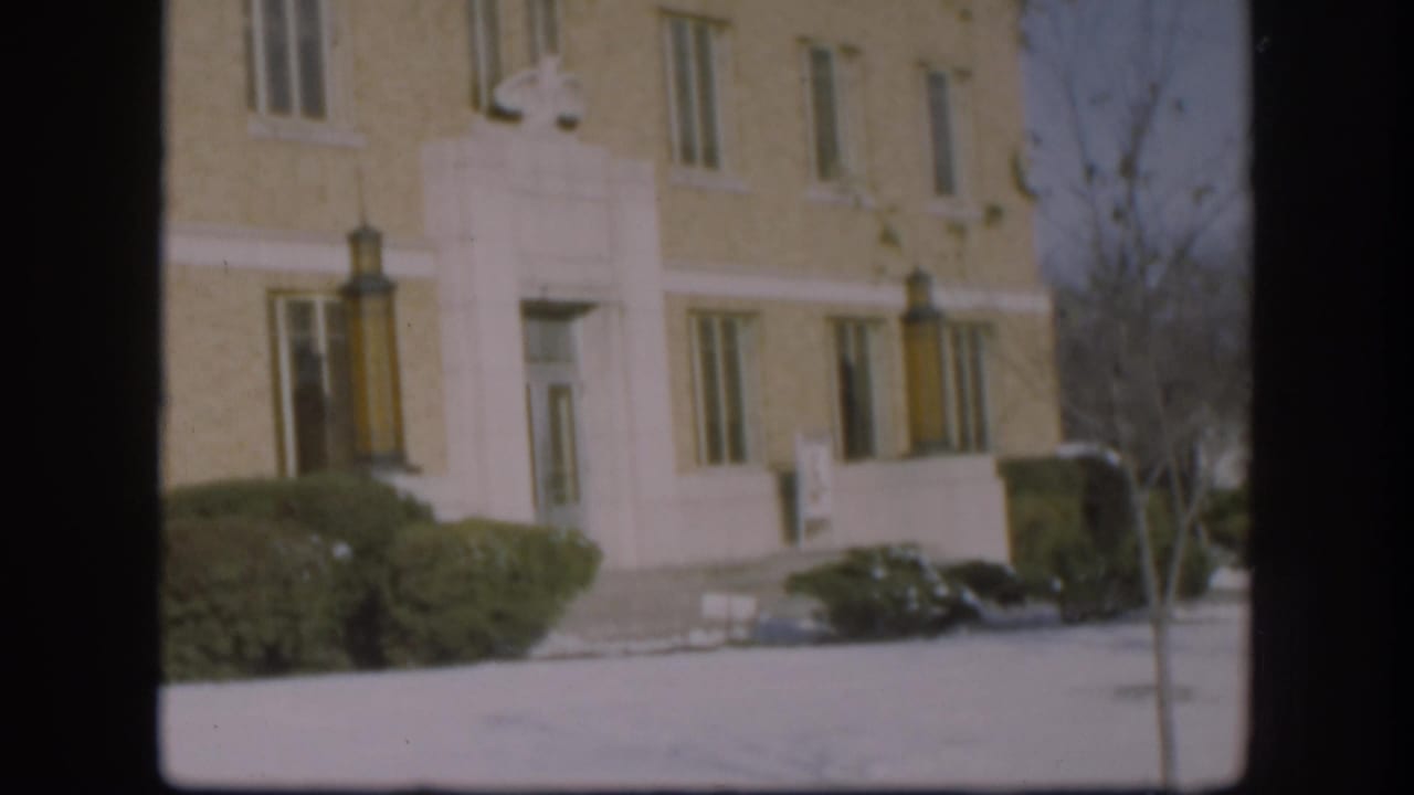 Front facade of a beige brick multi-storey building with white stone trim, snow-covered grounds, evergreen shrubs, and a bare tree at right.