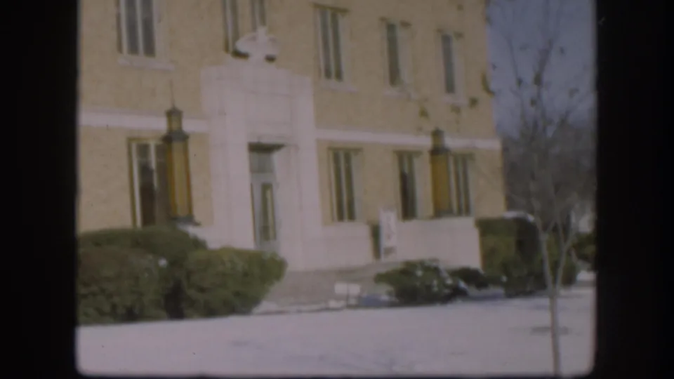 Wide shot of a beige brick courthouse facade with white stone classical detailing, snow-covered grounds and bare winter tree, Kansas circa 1958, 8mm color film.