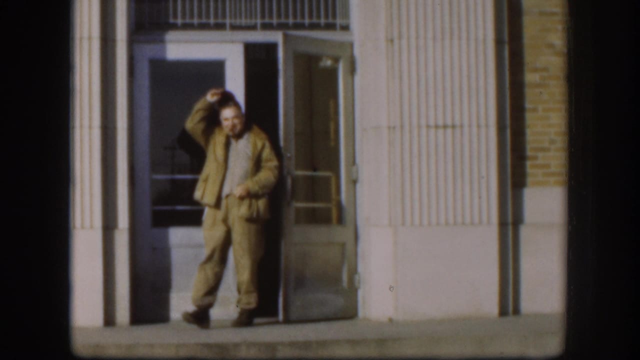 Man in tan two-piece suit standing at building entrance, hand raised to his hat, smiling toward camera.