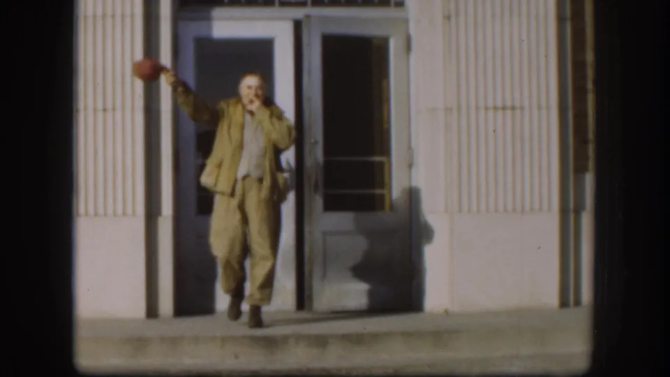 Man in tan suit on the steps of an institutional building, waving his hat overhead in greeting toward the camera, Kansas, circa 1958 — 8mm color footage.