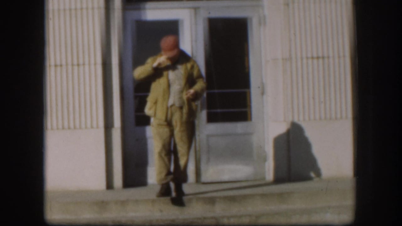 Middle-aged man in red cap and khaki jacket descends institutional building steps in what appears to be 1958 Kansas; glass doors and ribbed facade visible behind him.