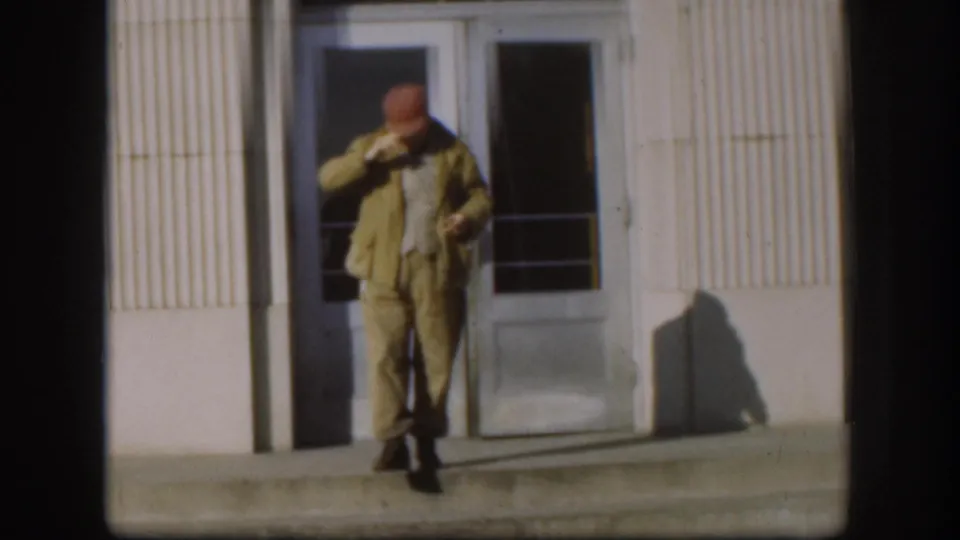 Middle-aged man in red cap and khaki jacket descends institutional building steps in what appears to be 1958 Kansas; glass doors and ribbed facade visible behind him.