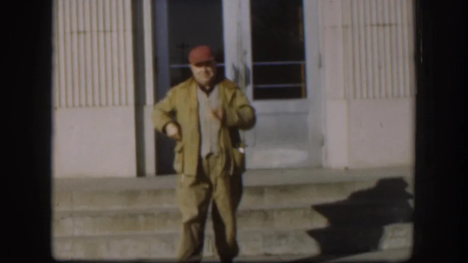 Man in red cap and khaki jacket walks away from building steps toward camera; concrete steps and ribbed facade visible behind him in mid-1950s color footage.