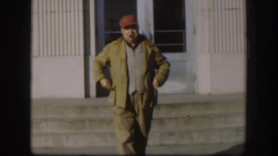 Close-medium view of man in red cap and tan jacket facing camera; mid-century institutional building facade and window visible behind him in 1958 Kansas.