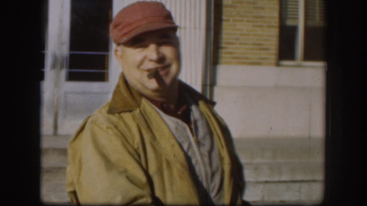 Medium close-up of man in tan jacket and red cap with a cigar in his mouth, smiling, brick building behind him