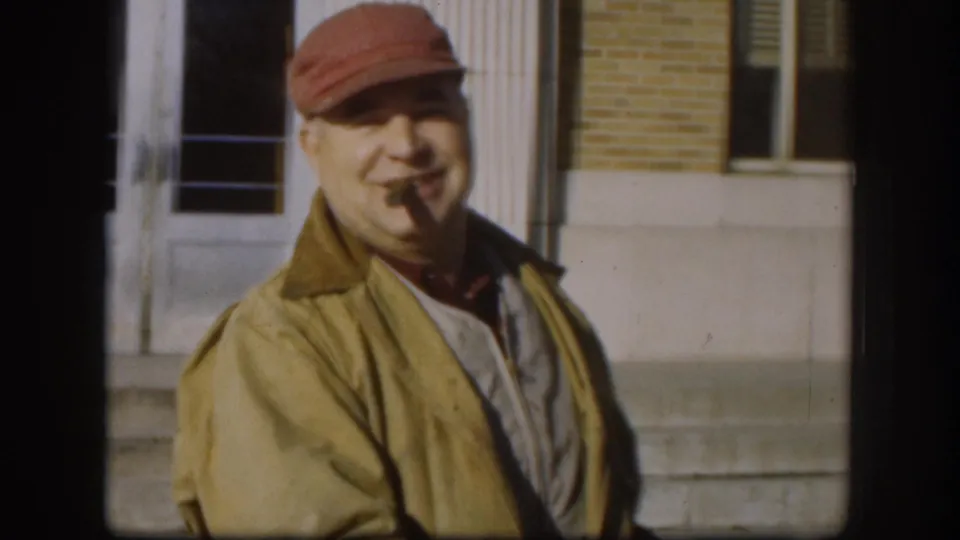 Medium shot of a man in an olive jacket and red cap with cigar in mouth, smiling outside a brick building, Kansas circa 1958