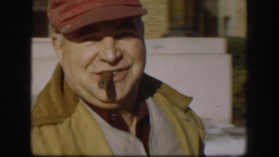 Close-up of a middle-aged man in a red peaked cap and olive jacket grinning with a cigar in his mouth, circa 1958 8mm home movie