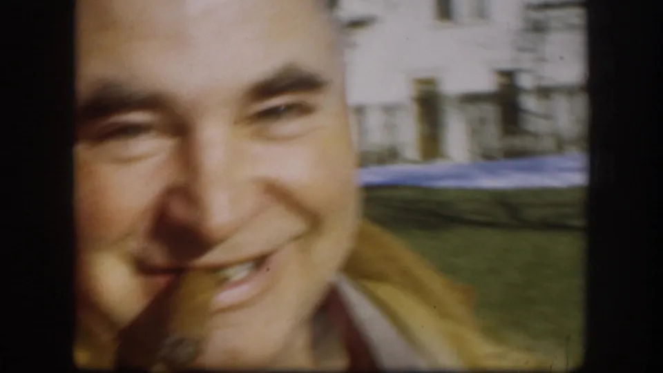 Extreme close-up of a middle-aged man laughing broadly toward the camera, green lawn glimpsed behind him, late-1950s 8mm home movie